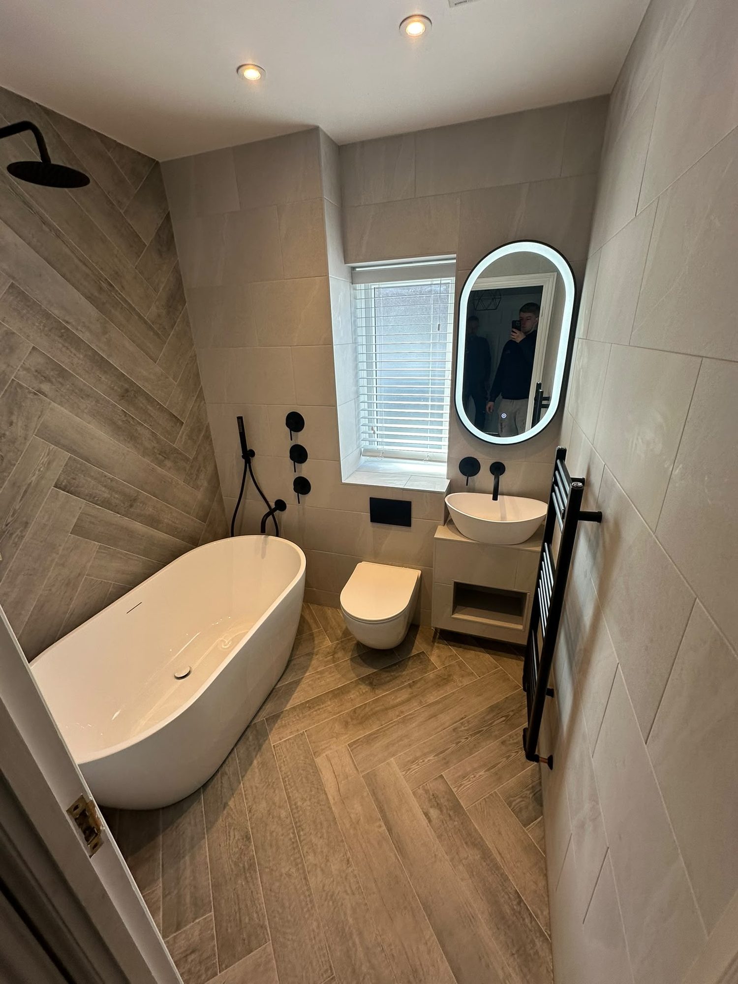 Freestanding bath with matt black taps, herringbone floor tiling and LED mirror by N Monk Plumbing & Heating