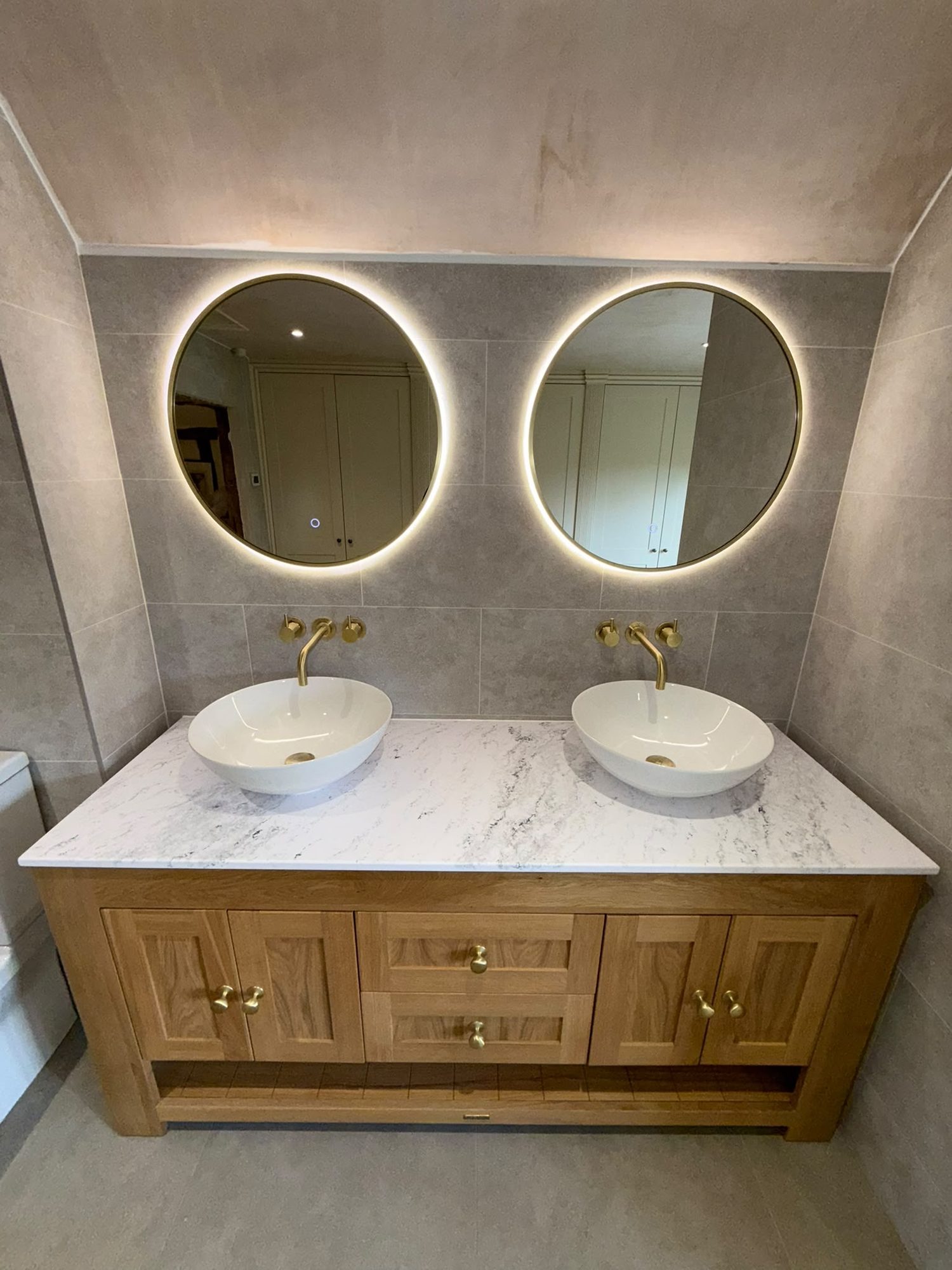 Twin countertop basins on a marble top with brass taps and backlit round mirrors by N Monk Plumbing & Heating